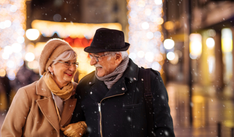 An older couple walking at night through a snow covered lit up city.